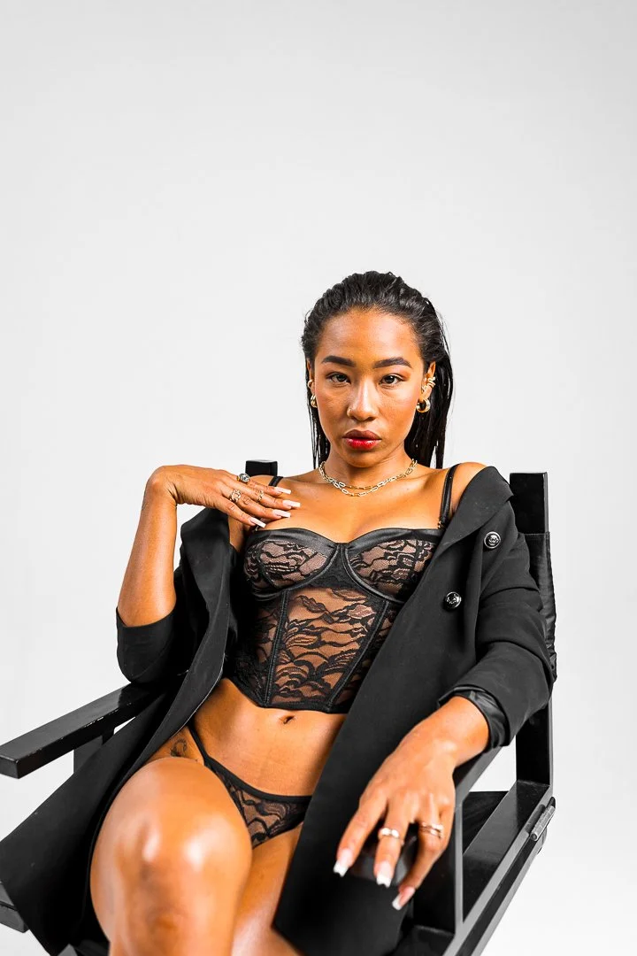 A woman sitting in a black chair, wearing black lace lingerie and a black blazer draped over her shoulders, with jewelry and polished nails, against a plain white background.