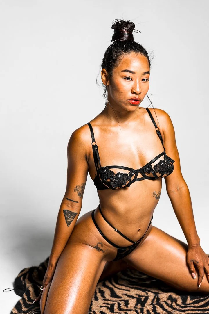 A woman with dark hair in a bun, wearing black lingerie with floral embroidery, sitting on a zebra-print rug against a plain white background.