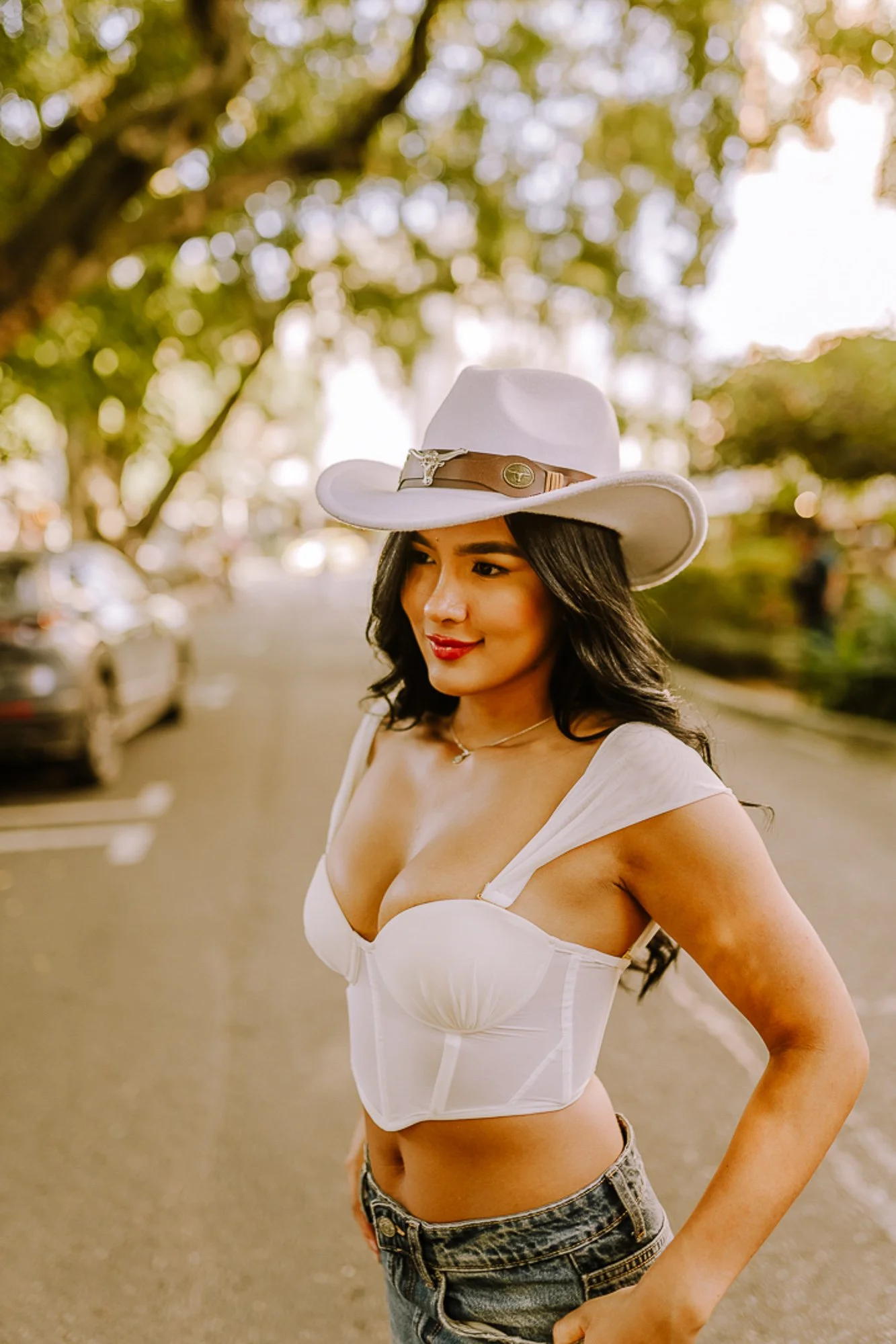 A woman with long black hair wearing a white cowboy hat, a white bustier top, and jeans, standing outdoors on a street with trees and cars in the background.