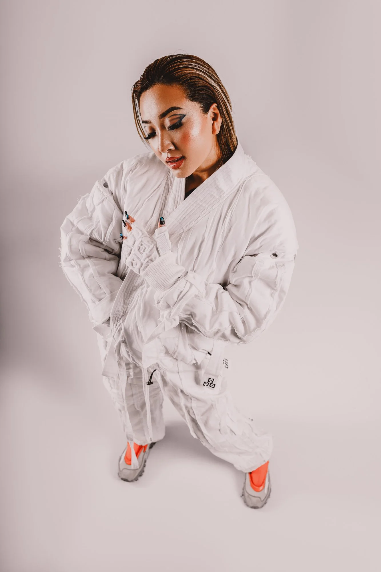 A woman in a white gi kneeling with her head slightly bowed, wearing colorful sneakers against a plain white background.