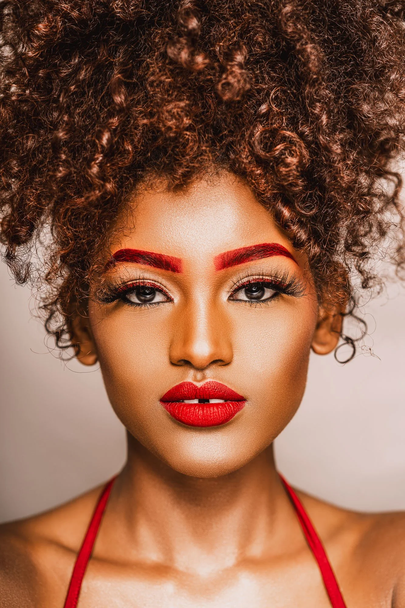 A woman with tan skin, curly hair, bold red eyebrows, and bright red lipstick. She is wearing makeup, including eye makeup with gold and black tones, and is dressed in a red strap top.