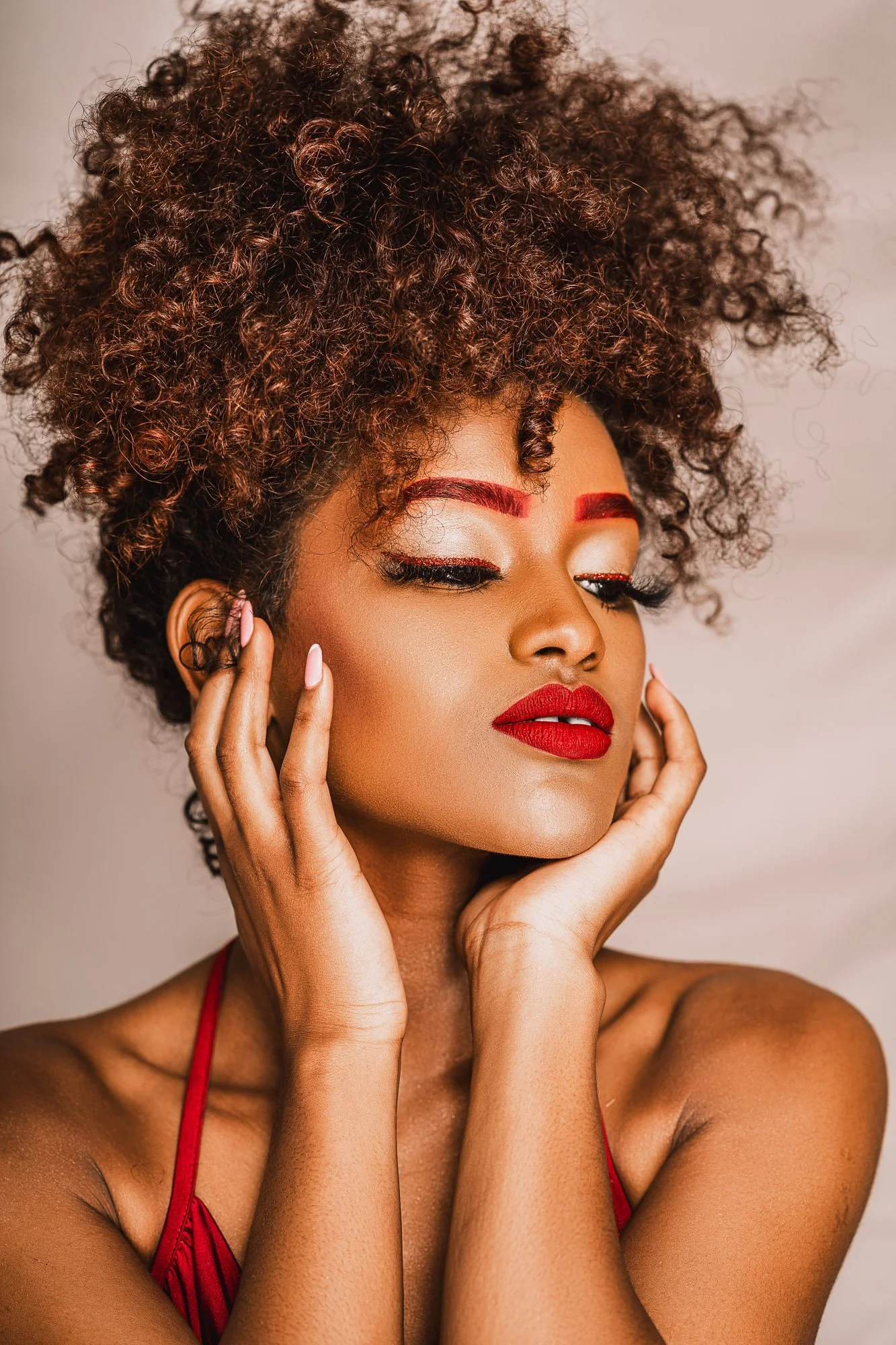 A woman with curly hair and vibrant makeup, including red eyebrows and lipstick, posing with her hands touching her face.