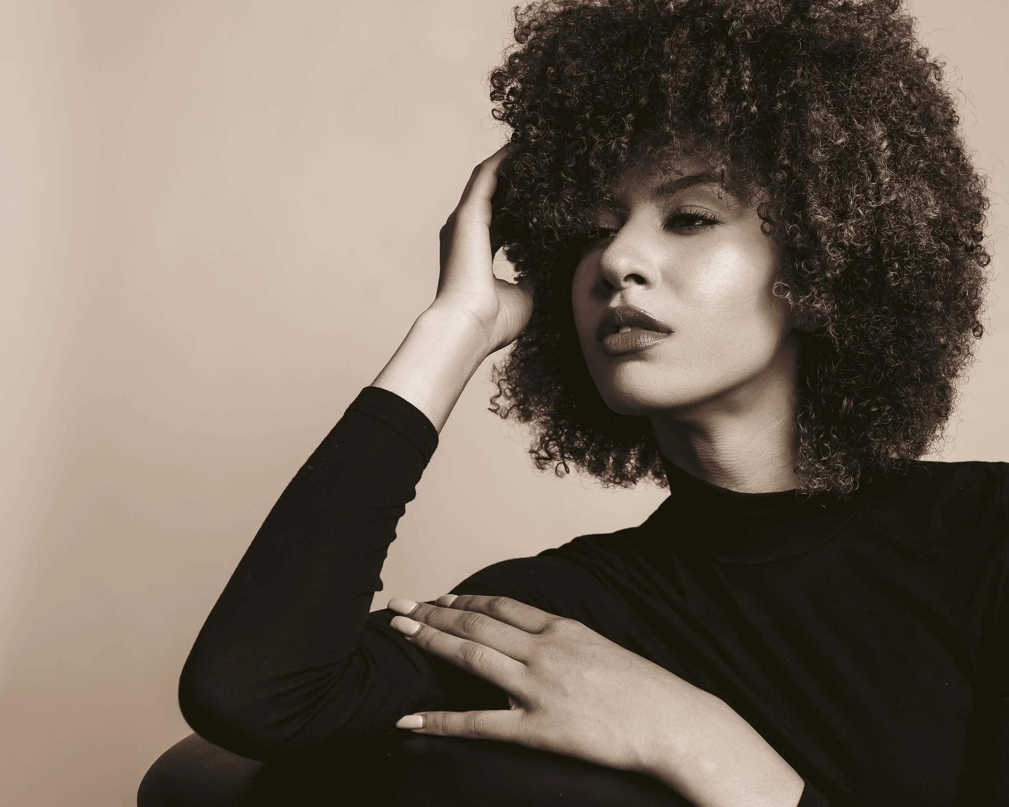 A woman with natural curly hair posing with her hand on her head and shoulder, looking confident, wearing a black long-sleeve top, in a studio setting with a neutral background.