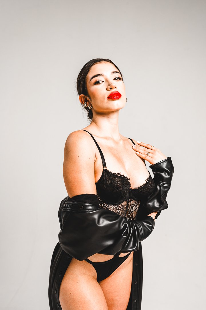 A woman with dark hair, bold red lipstick, and multiple earrings is wearing a black lace lingerie top, black underwear, and a black leather jacket draped over her shoulders. She has a confident pose against a plain, light-colored background.