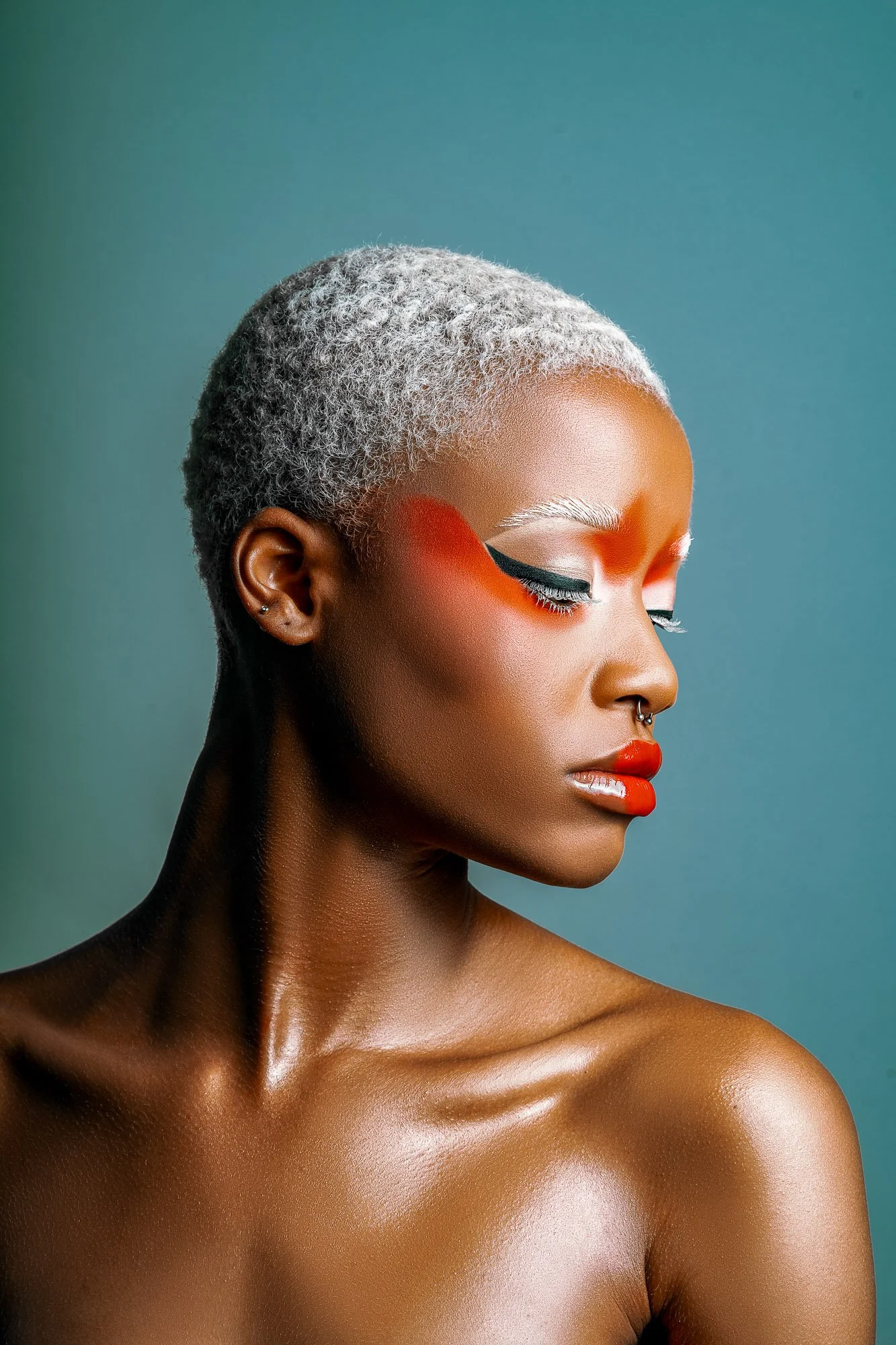 A woman with short gray hair wearing bold makeup, including orange eye shadow, white eyeliner, and red lipstick, with a septum piercing, posed against a muted teal background.