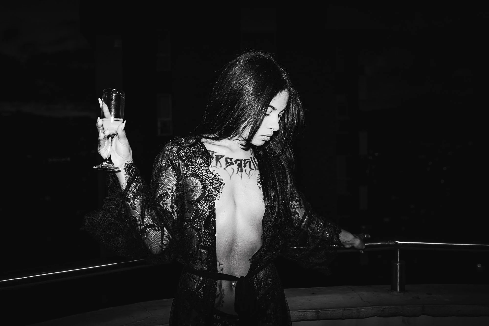 A woman with long dark hair, lace robe, and tattoos, holding a glass of champagne at night with a city skyline in the background.