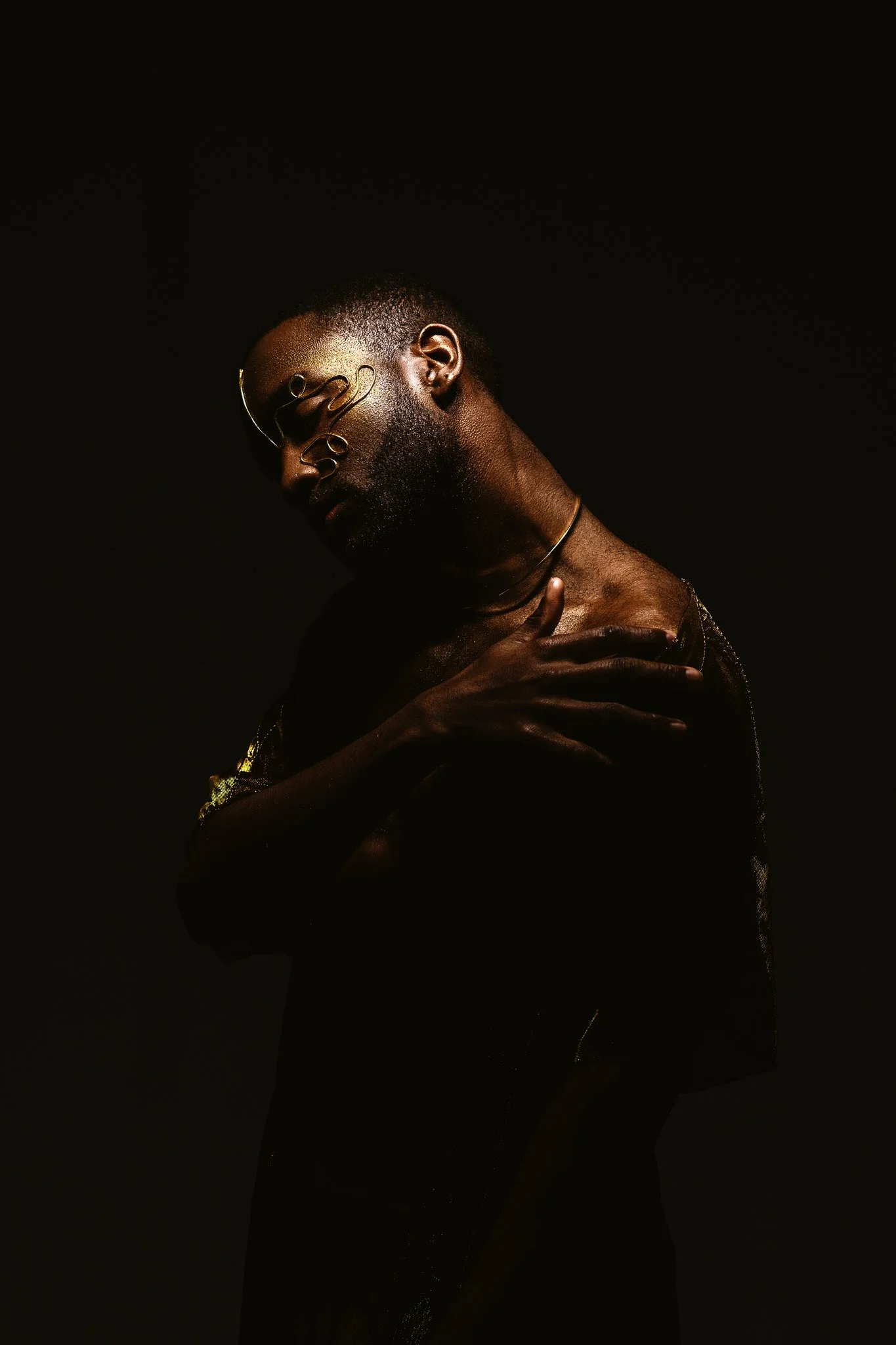 A dark-skinned person with short hair and a beard, wearing glasses and a choker, is posing against a black background with their head tilted to the side and hand on their chest.