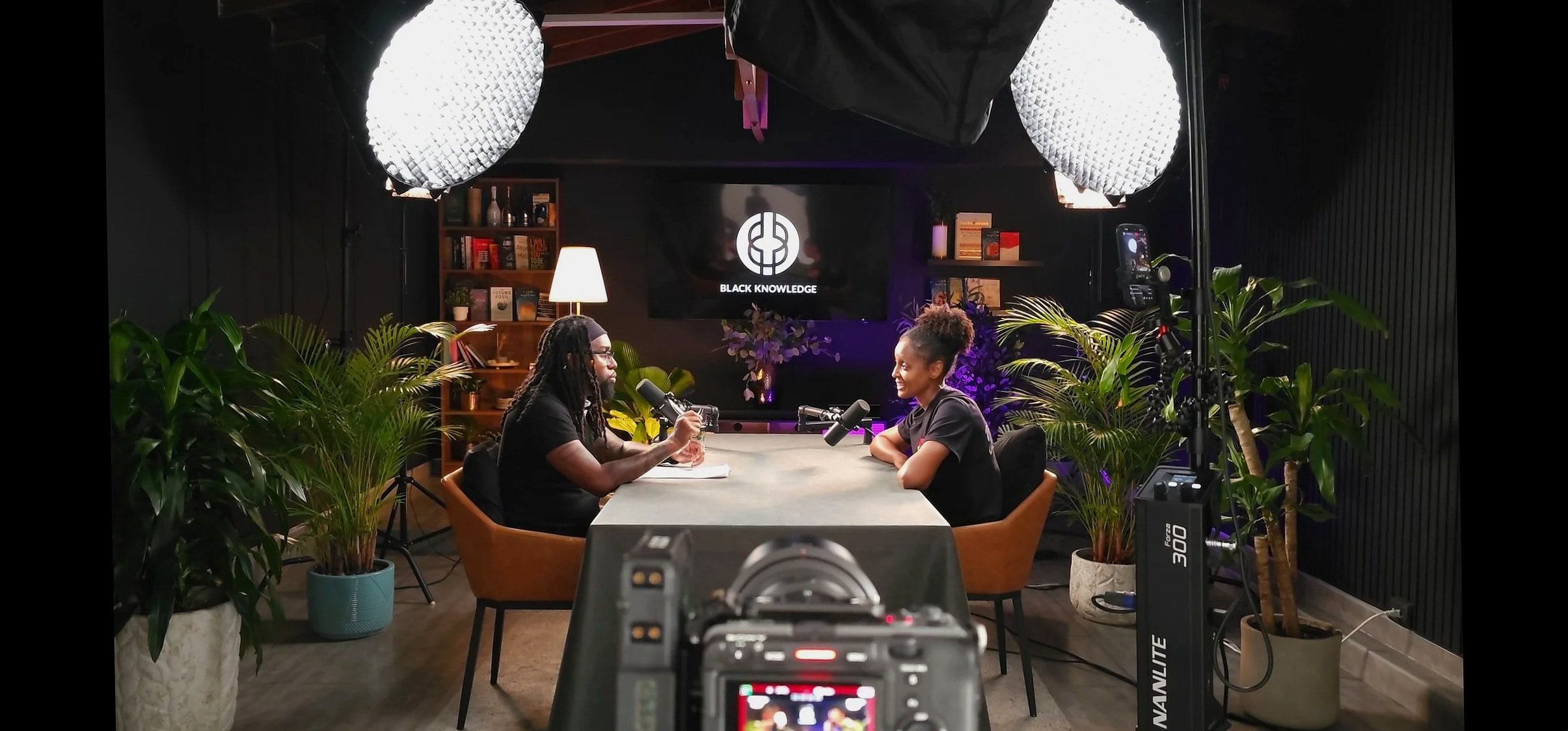 Two people sitting at a table in a recording studio, engaging in a conversation with microphones in front of them. The background displays a screen with the logo 'Black Knowledge' and a bookshelf. Studio lighting equipment is visible, and there are green plants around the setting.