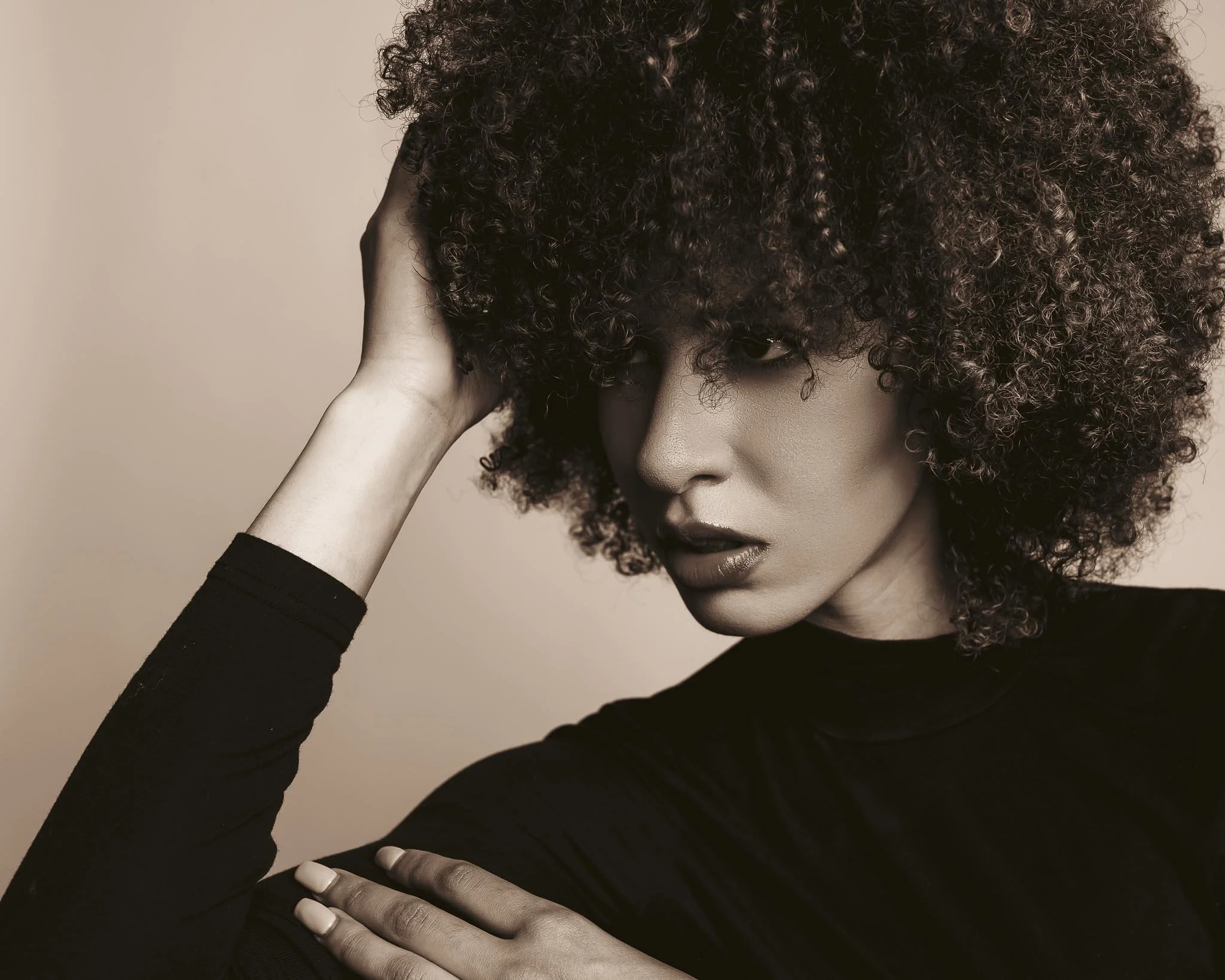 A Black woman with voluminous curly hair, wearing a black top, posing with her hand resting on her head and looking thoughtfully to the side.