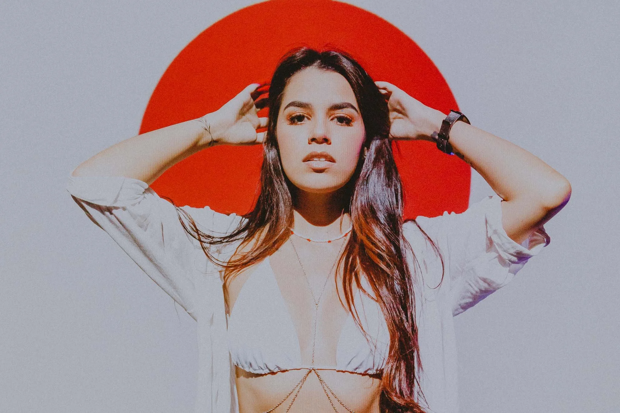 A young woman with long dark hair posing against a white background with a large red circle behind her head, wearing a white crop top and accessories.