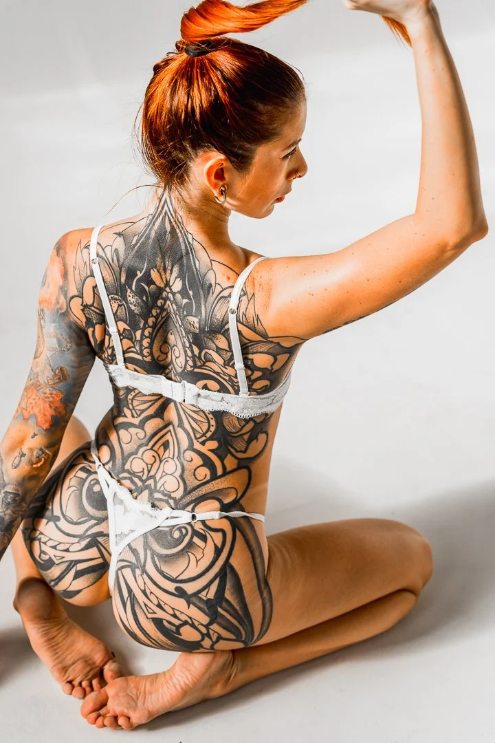 A woman with orange hair styled in a bun, tattooed arms and back, wearing white lingerie, kneeling on the floor, flexing her right arm.
