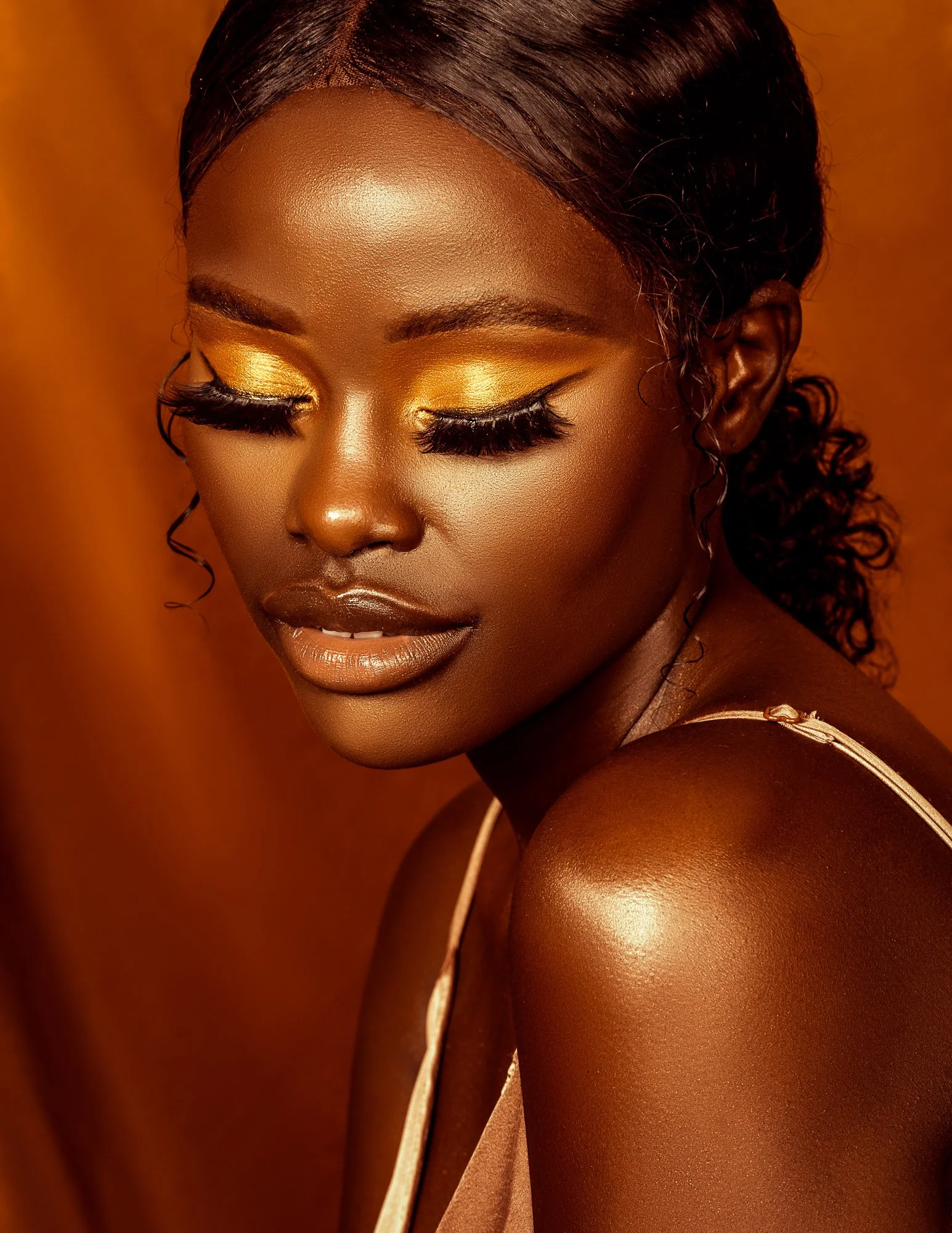 Close-up of a woman with dark skin and curly hair, wearing gold eyeshadow and lipstick, with her eyes closed and a soft smile, against a warm brown background.
