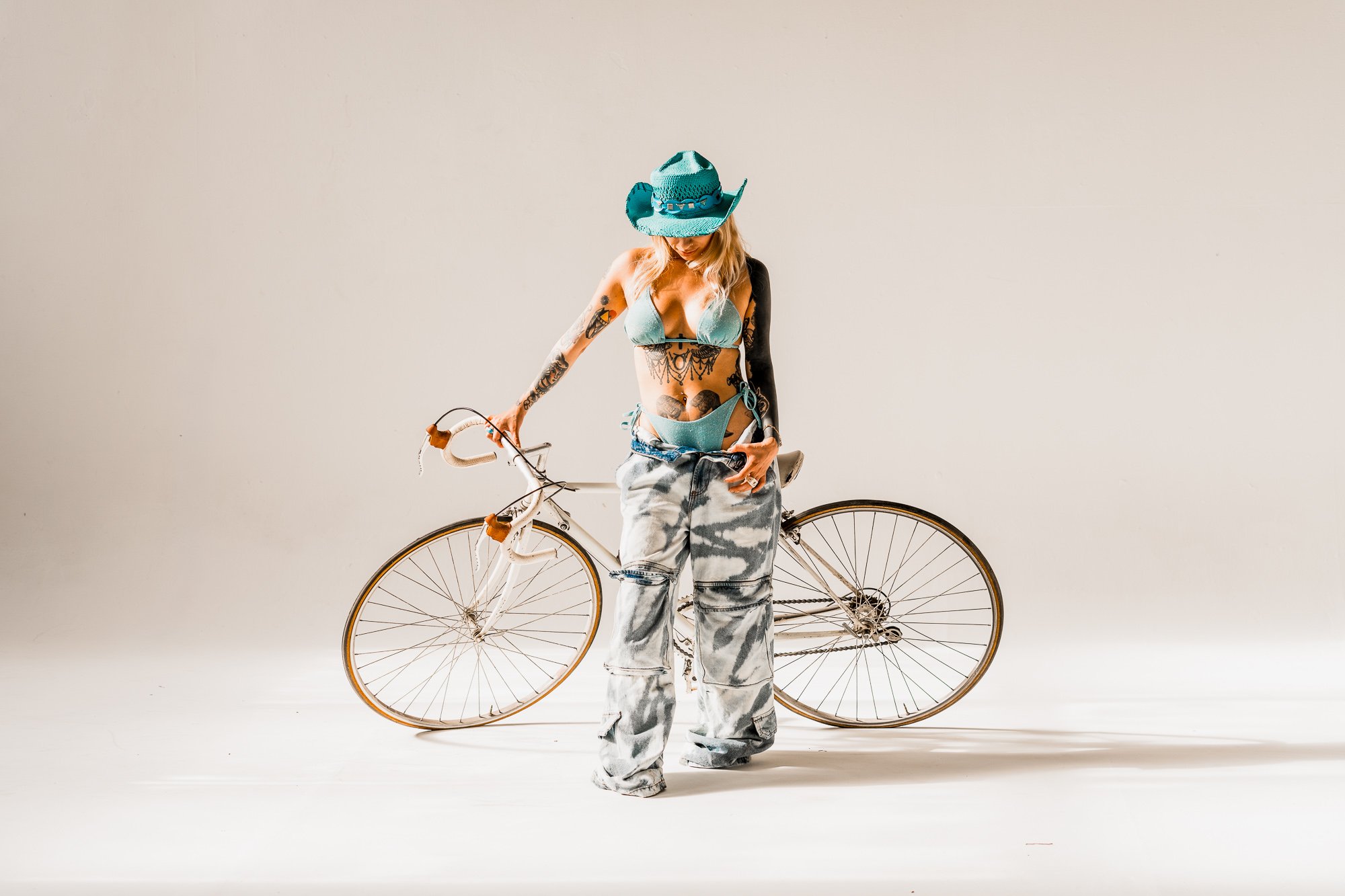 Woman wearing a blue hat and bikini top, standing with a bicycle against a plain light-colored background.