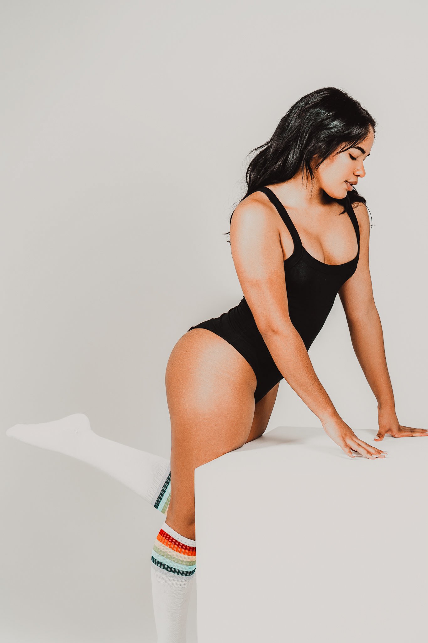 A woman with long dark hair wearing a black tank top and rainbow-striped knee-high socks. She is on her hands and knees, with one leg lifted and bent at the knee, against a plain white background.