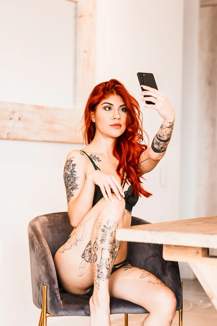 A young woman with red hair and tattoos sitting on a gray chair in a room, taking a selfie with her phone.