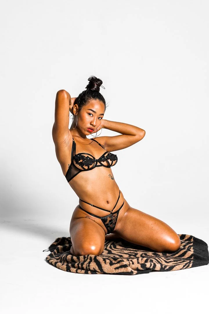 A woman kneeling on a zebra print rug, posing in black lace lingerie, with her hands behind her head, in front of a plain white background.