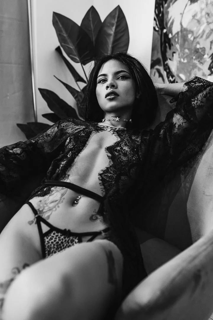 A woman with dark hair and tattoos wearing black lace lingerie and a choker, reclining on a sofa with large plants and abstract artwork in the background.