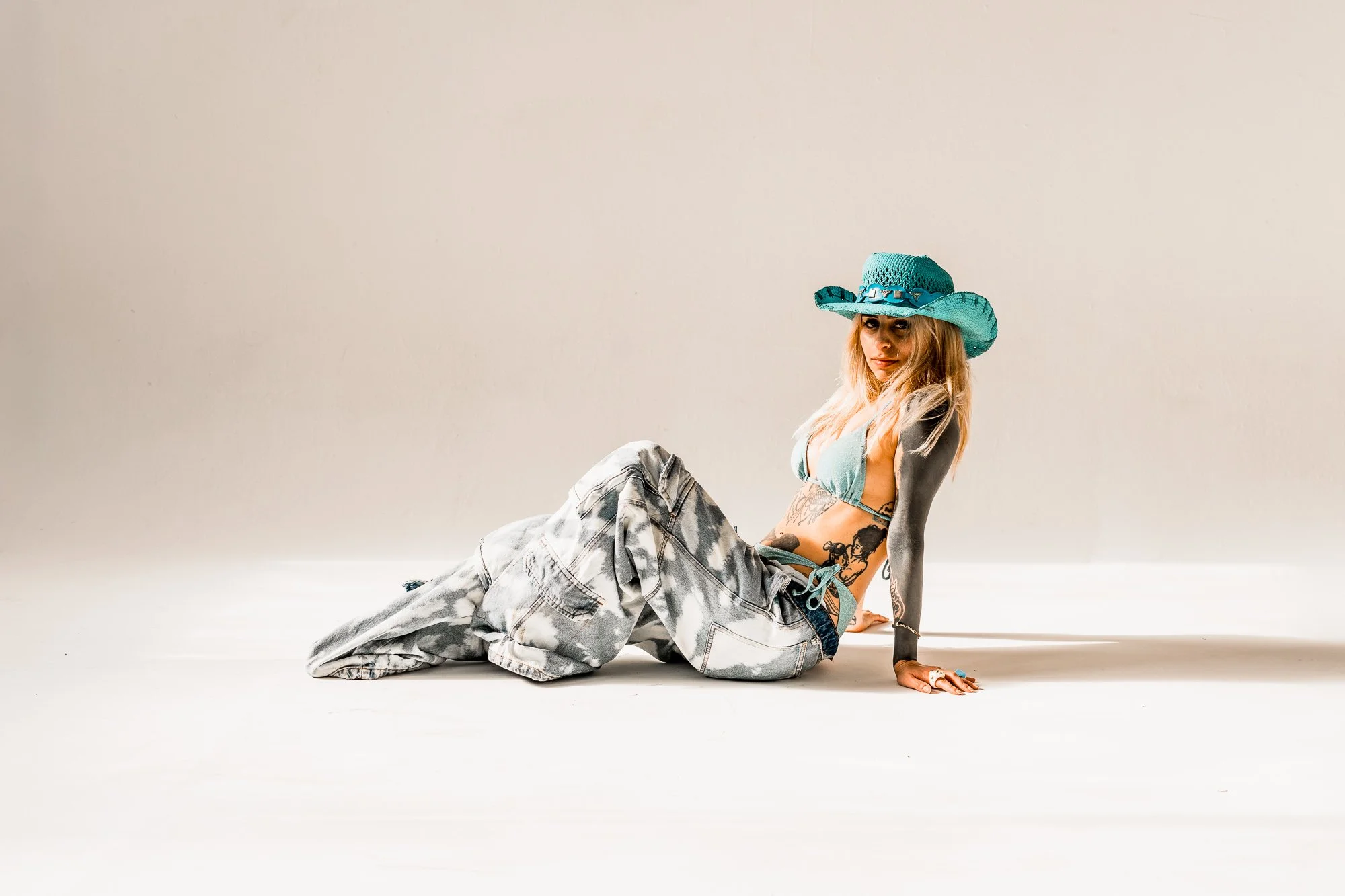 Woman sitting on the floor wearing a large turquoise hat, bikini top, baggy acid-wash jeans, and multiple rings and tattoos.