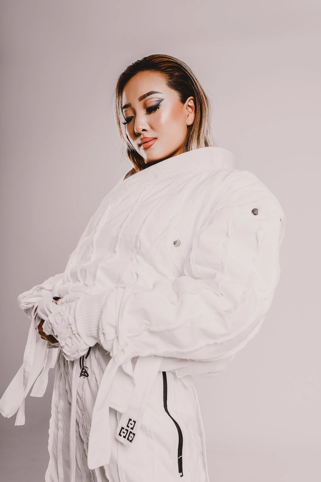 A woman wearing a white quilted jacket with a high collar and long sleeves, standing against a plain light background, with makeup including winged eyeliner and lip color, and brown hair styled back.