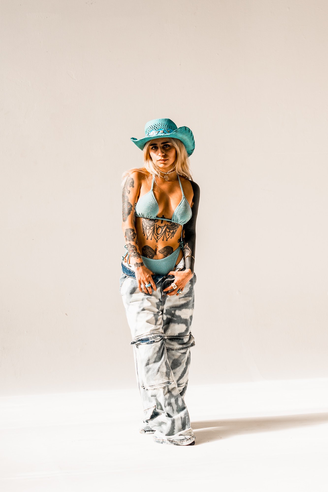 A woman wearing a large turquoise hat, bikini top and bottom, and baggy camouflage pants, standing against a plain light background.