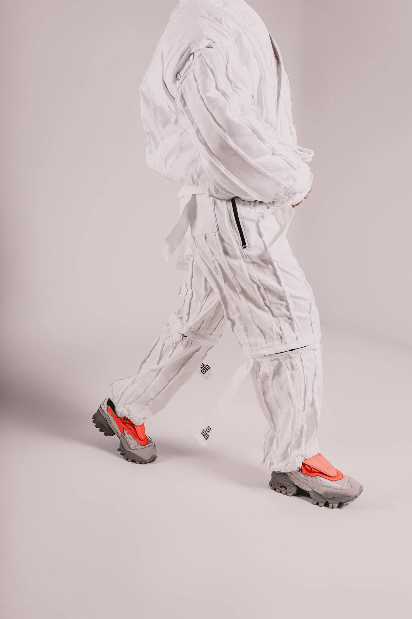 Person wearing white puffer jacket, baggy white pants, and gray and orange sneakers against a neutral background.