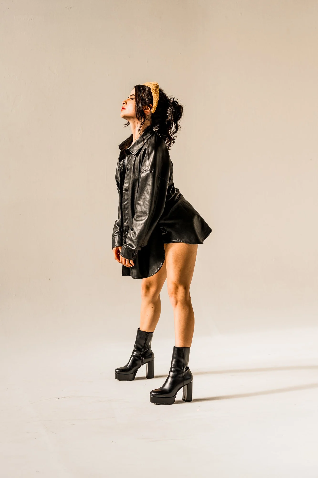A woman standing against a plain light-colored background, wearing a black leather jacket, black high-heeled boots, and a small yellow headband. She has dark wavy hair and is shown in profile with her eyes closed.