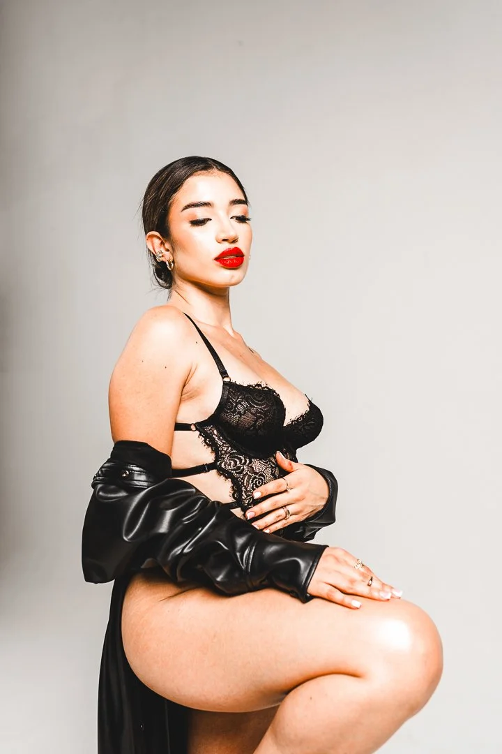 A woman with dark hair and red lipstick sitting with her eyes closed, wearing black lace lingerie and a black leather jacket draped over her shoulders.