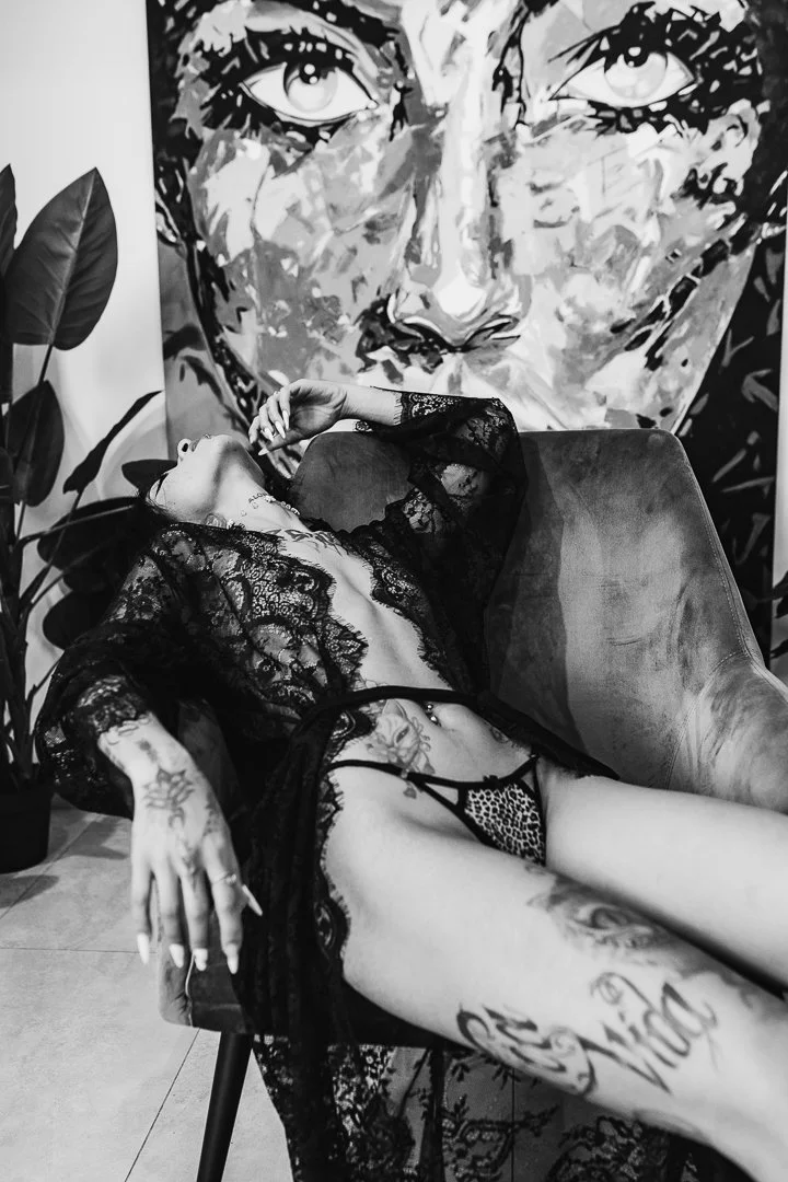 A woman with tattoos and dressed in lace lingerie, reclining on a chair with her head back and eyes closed, in front of a large black and white face mural.