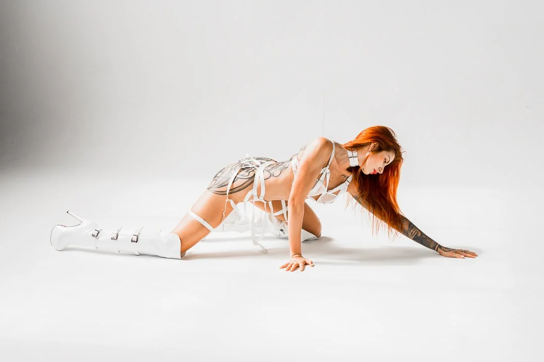 A woman with long red hair and tattoos on her arm and leg is on all fours on a white background, dressed in white strappy clothing and white high-heeled boots.