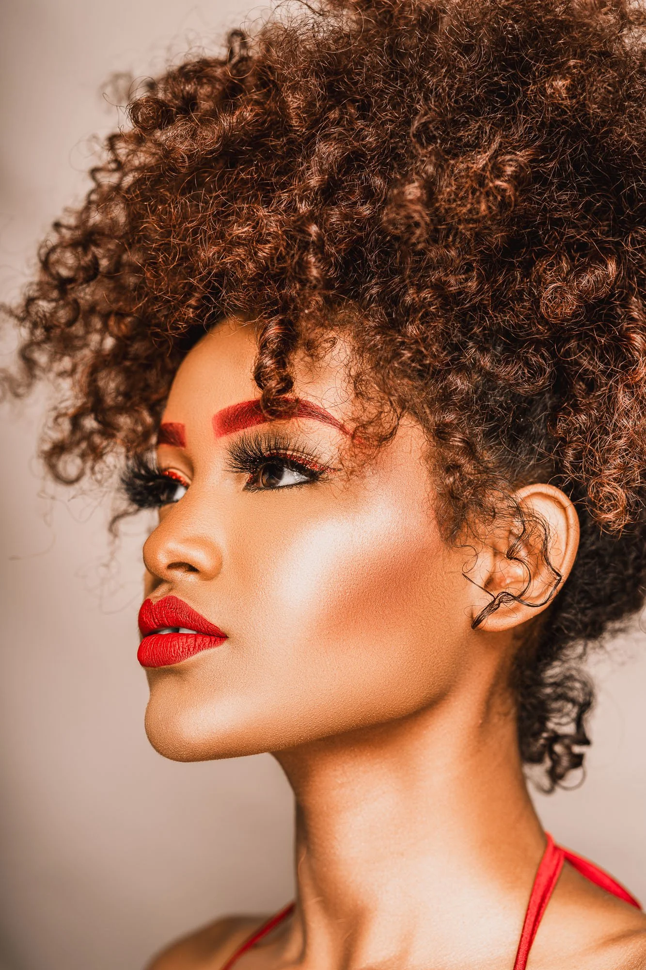A woman with curly brown hair and red makeup, including bold red eyebrows and lipstick, is looking to the side.