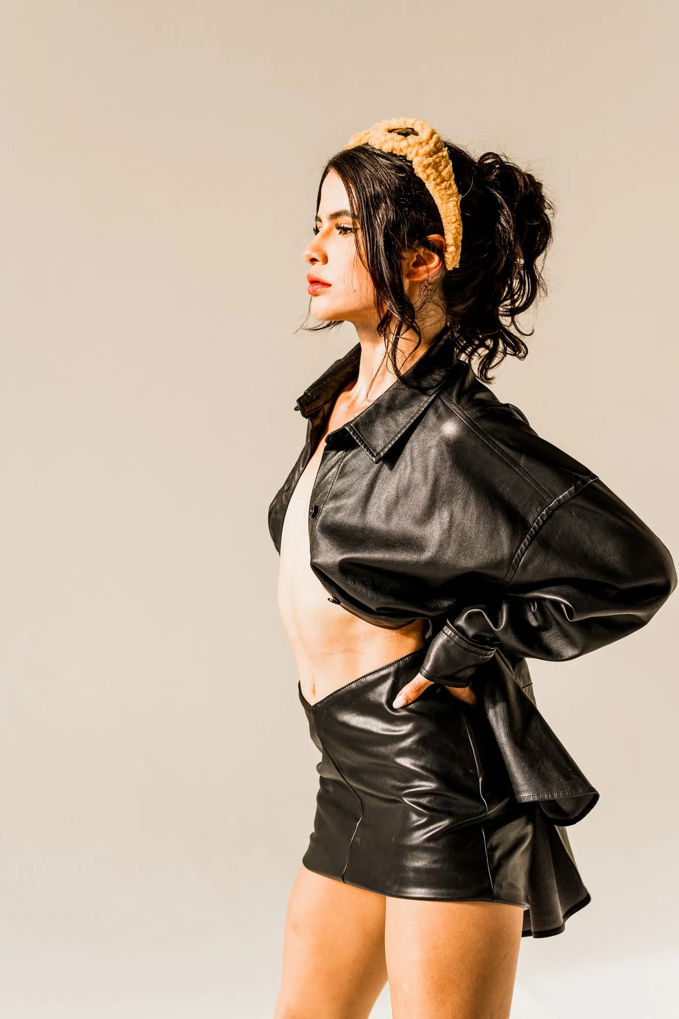 A woman with curly dark hair wearing a beige headband, black leather jacket, and matching black leather skirt against a neutral background.