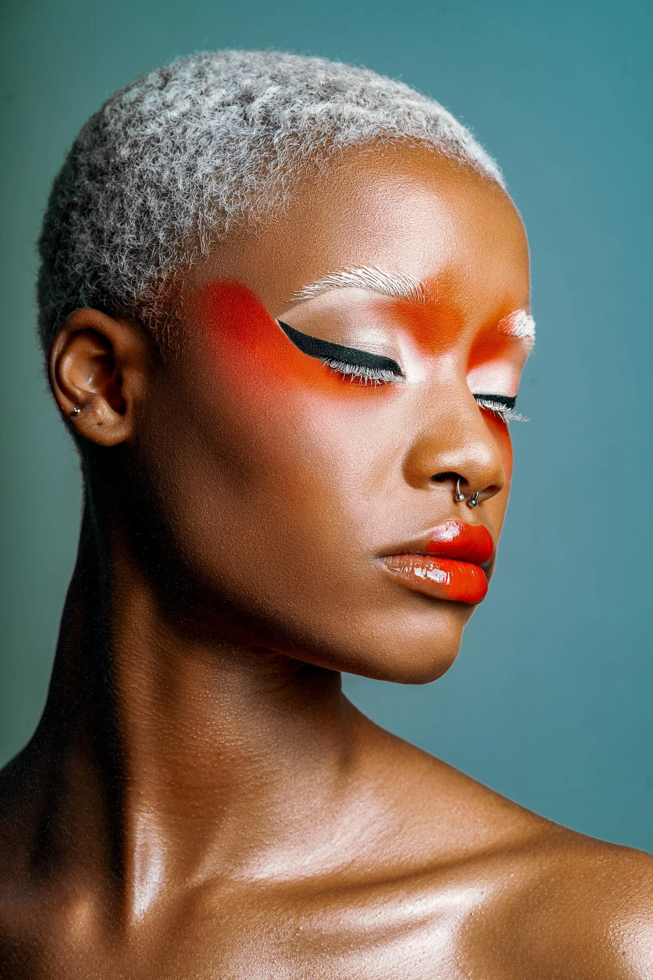 Close-up of a woman with short platinum blonde hair, wearing bold red lipstick, dramatic eye makeup with white and black eyeliner, and a septum piercing.