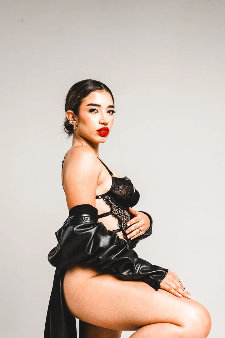 A woman with dark hair tied back, wearing red lipstick, a black lacy lingerie top, and a black leather jacket draped over her shoulder, posing against a plain, light-colored background.