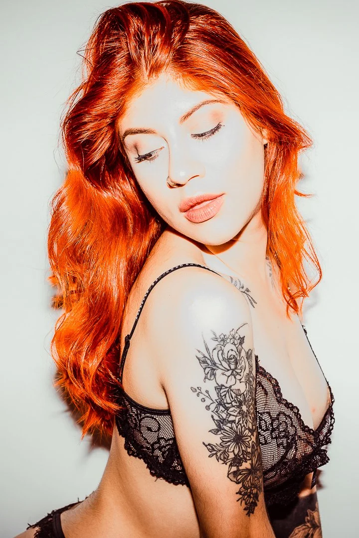 A woman with long, wavy red hair and tattoos on her shoulder, wearing black lace lingerie, is posing with her eyes closed and head tilted slightly downward against a plain background.
