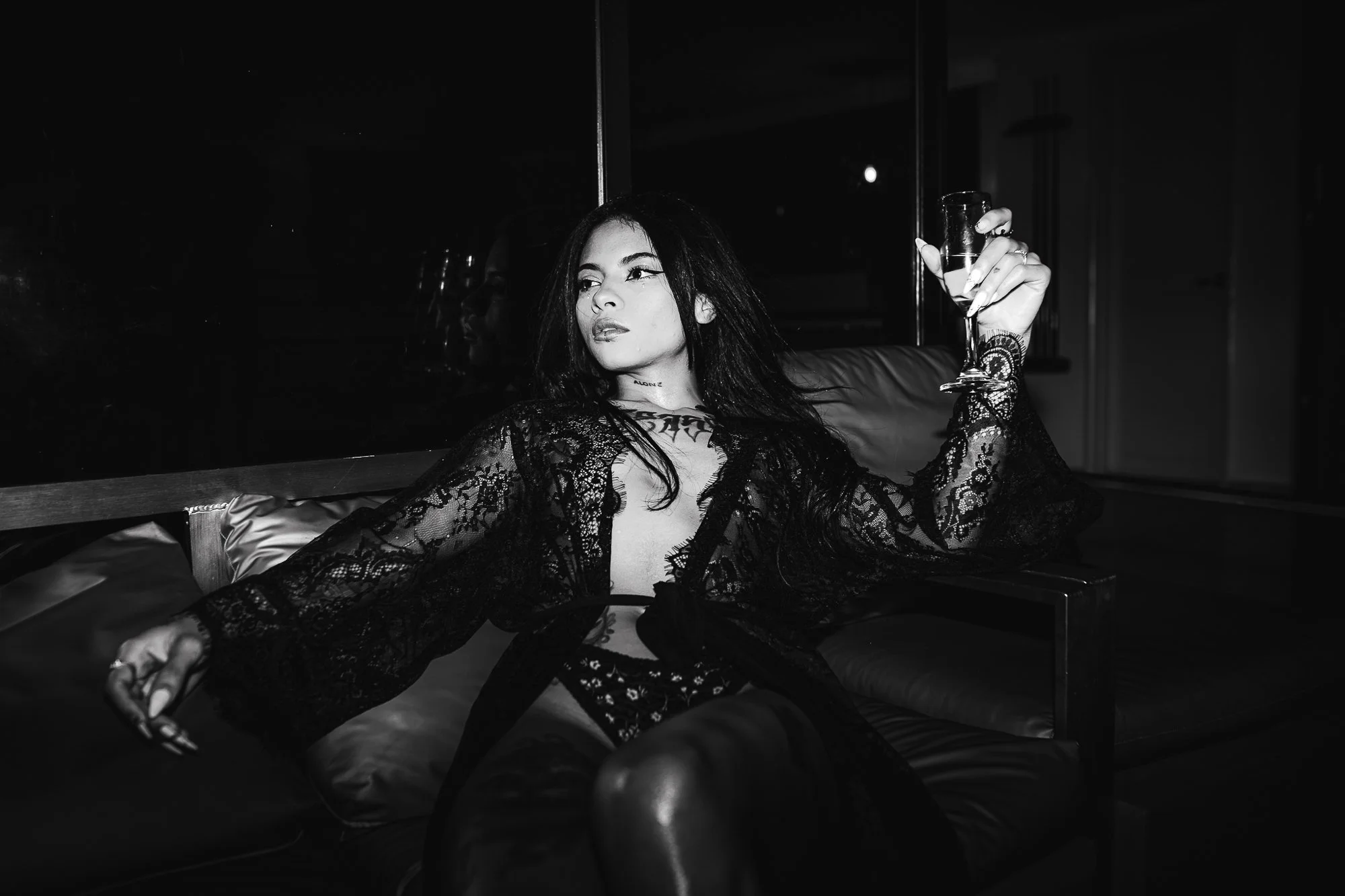 A woman in a black lace dress sits on a leather couch in a dimly lit room, holding a glass of wine in her right hand, with her head turned slightly to the side and a contemplative expression.