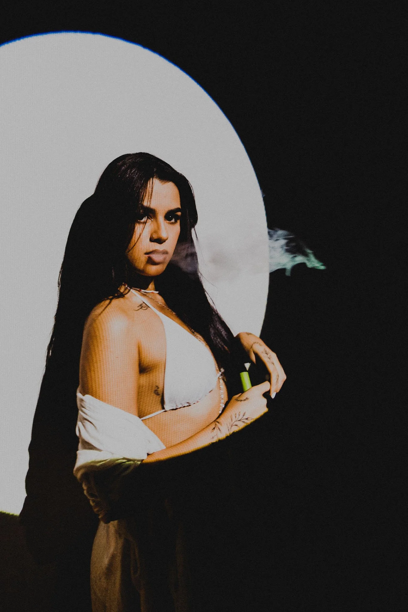 A woman with long dark hair, tattoos, and makeup, standing in front of a white circle against a dark background, holding a vape device and exhaling vapor.