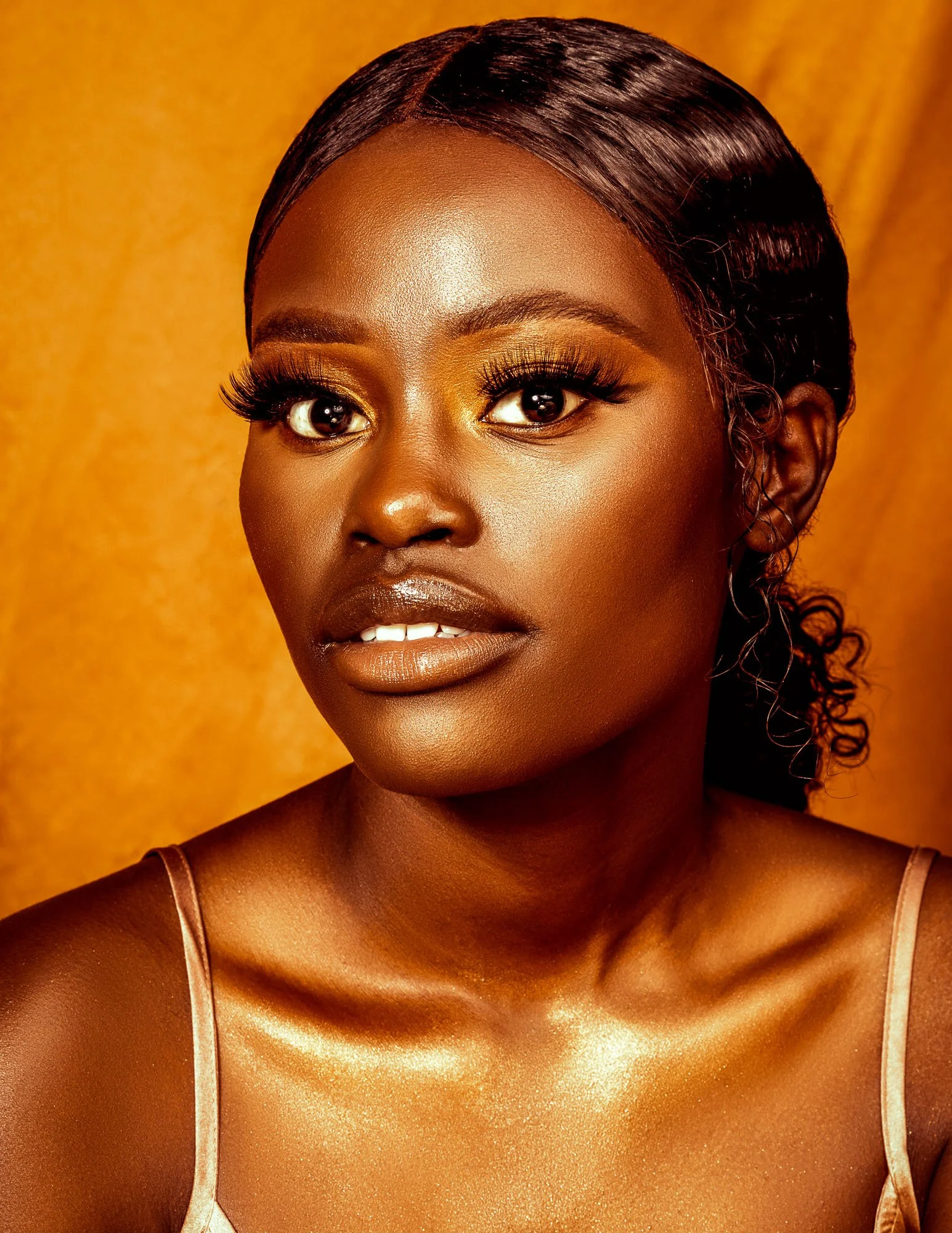 Close-up portrait of a woman with dark skin, makeup, and styled hair, wearing a satin camisole, against a gold background.