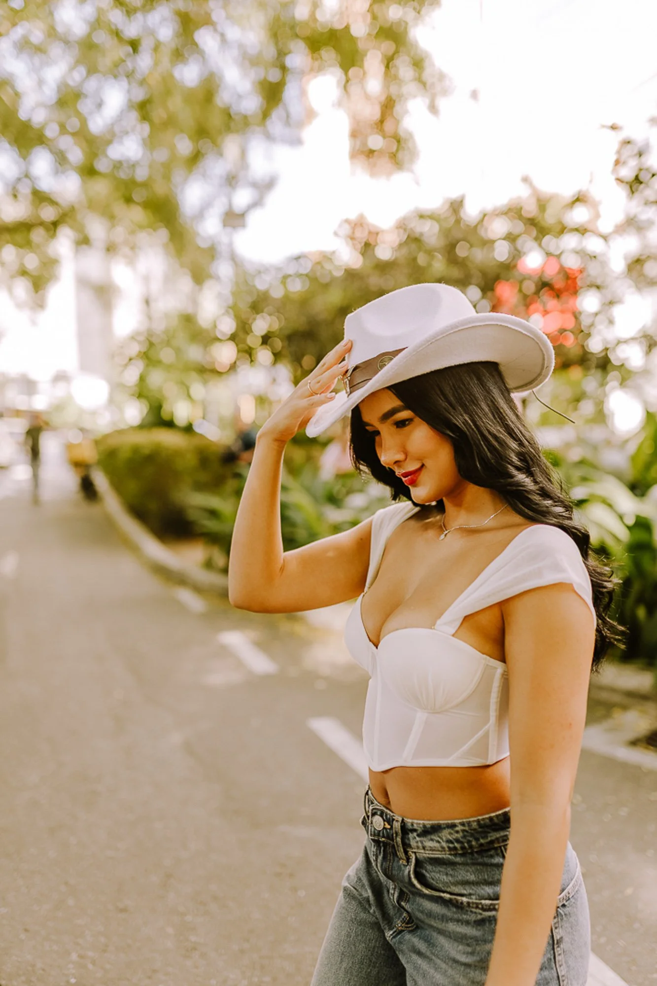A woman with long black hair in a white corset top, gray jeans, and a white hat standing outdoors on a tree-lined street, smiling and adjusting her hat.