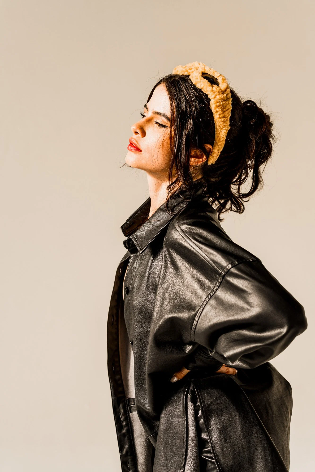 A woman with dark curly hair wearing a beige headband, a black leather jacket, and red lipstick. She is shown in profile with her eyes closed, standing against a plain background.