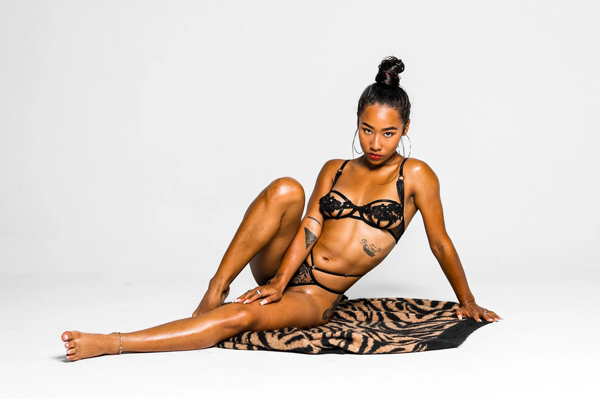 A woman with dark hair in a top knot hairdo, wearing black lingerie, sitting on a tiger-patterned rug against a plain white background.
