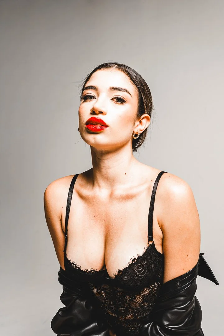 A woman with dark hair styled back, wearing red lipstick, a black lace top, and a black leather jacket off her shoulders, with a neutral background.
