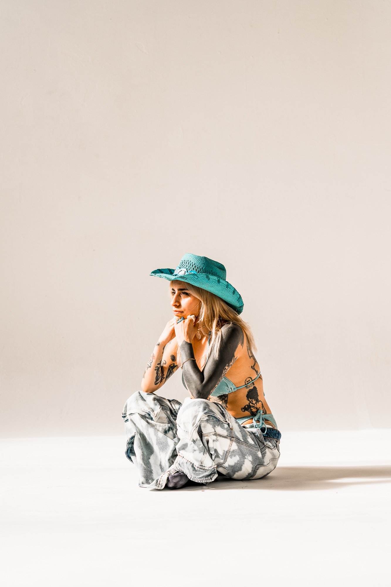 A woman sitting cross-legged on the floor, wearing a turquoise wide-brimmed hat, a bikini top, and loose tie-dye jeans with tattoos visible on her arms.