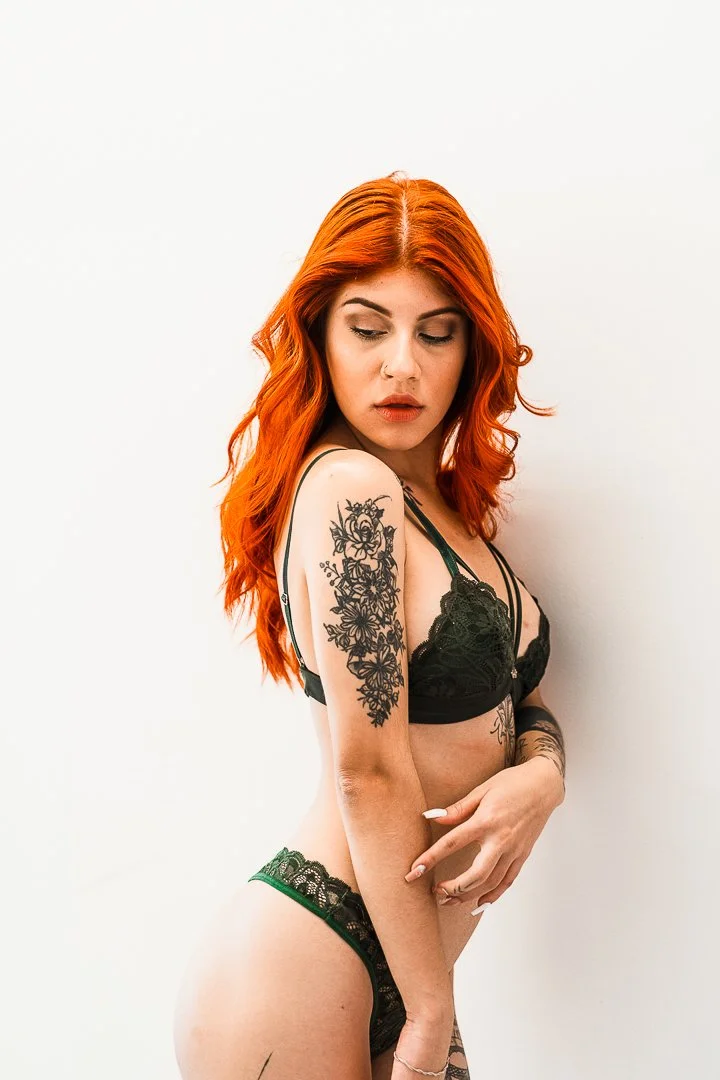 A woman with vibrant red hair, wearing black lace lingerie, standing against a plain white background.