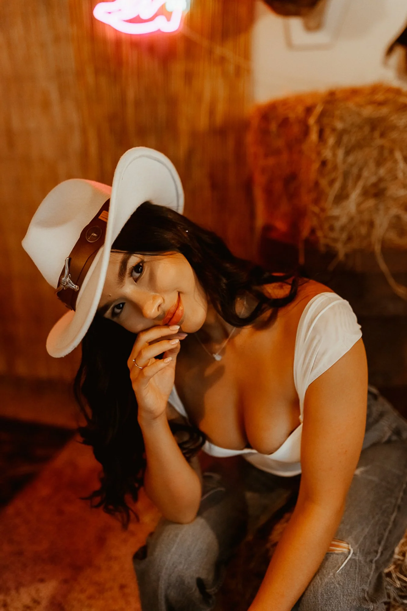 A woman with long dark hair wearing a white hat with a brown band, a white top, and gray jeans, posing indoors in a dimly lit setting with warm wood tones.