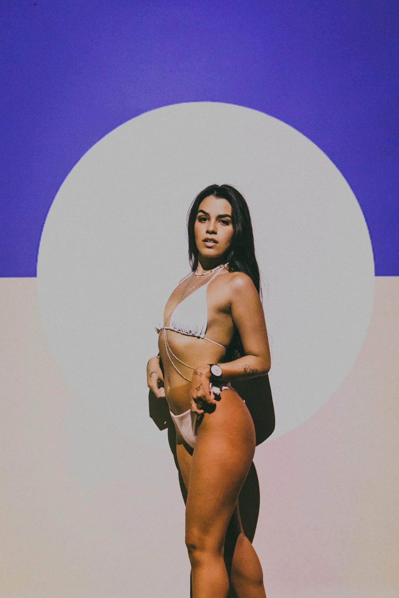 A woman in a white bikini with chains stands in front of a colorful background with a large circle and purple, white, and beige sections.