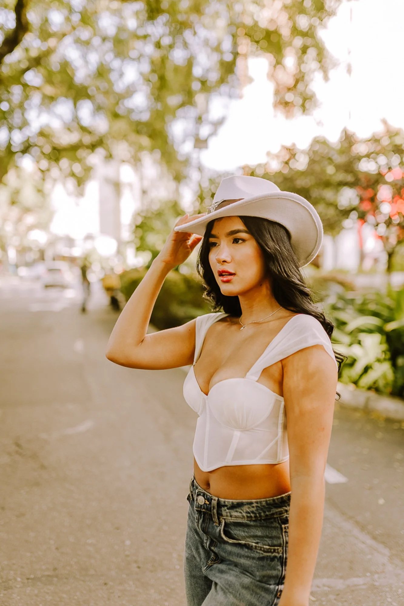 A young woman with black hair wearing a white hat, white top, and jeans standing outdoors on a tree-lined street.
