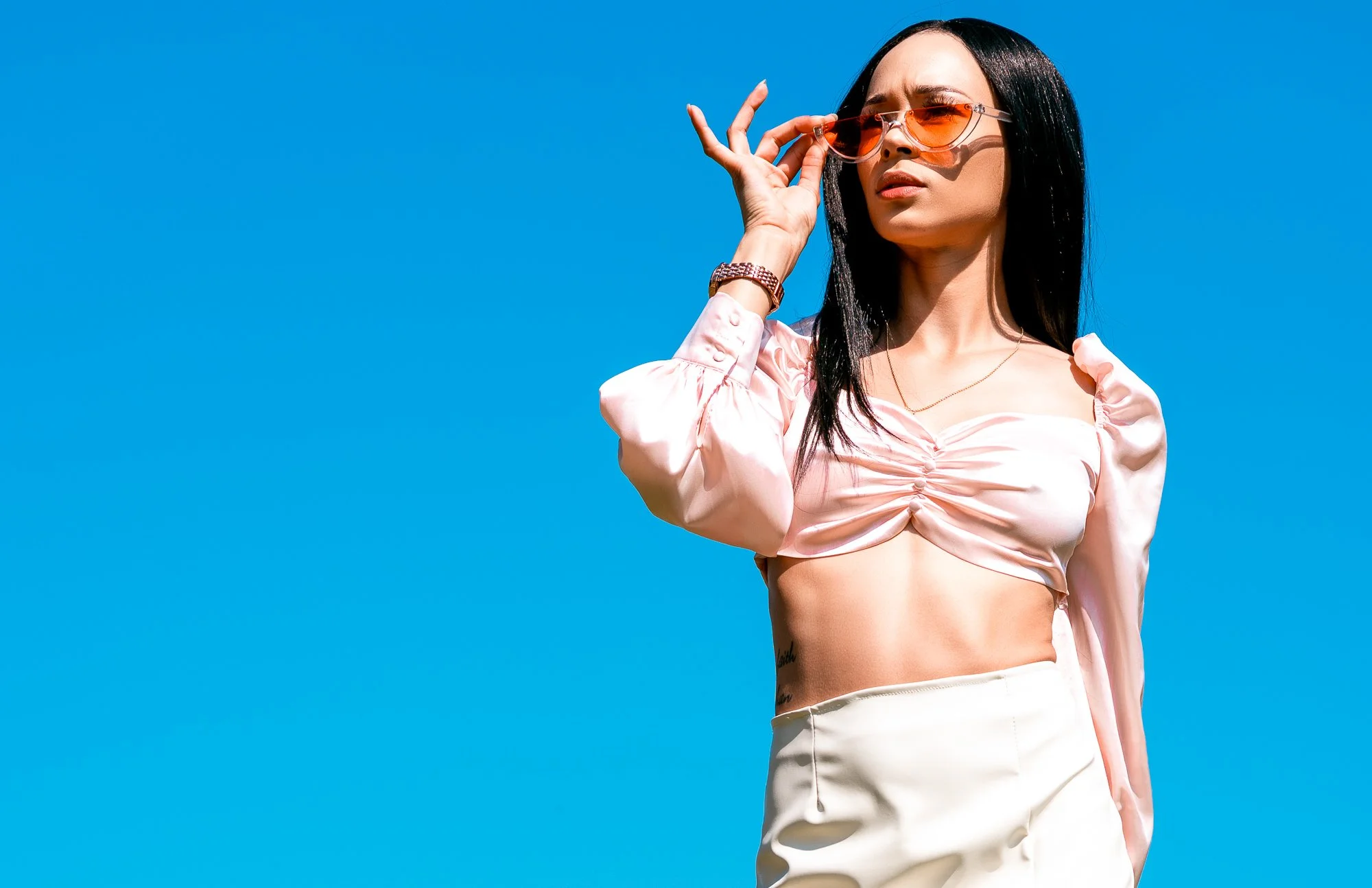 A stylish woman with long black hair wearing orange-tinted sunglasses, a pink cropped puff-sleeve top, and beige pants, holding her sunglasses with a blue sky in the background.