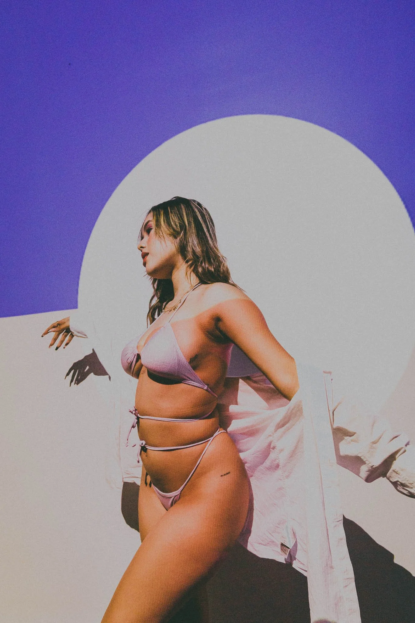 A woman wearing a light pink lingerie set and a white shirt poses against a multicolored background with a circular white shape.