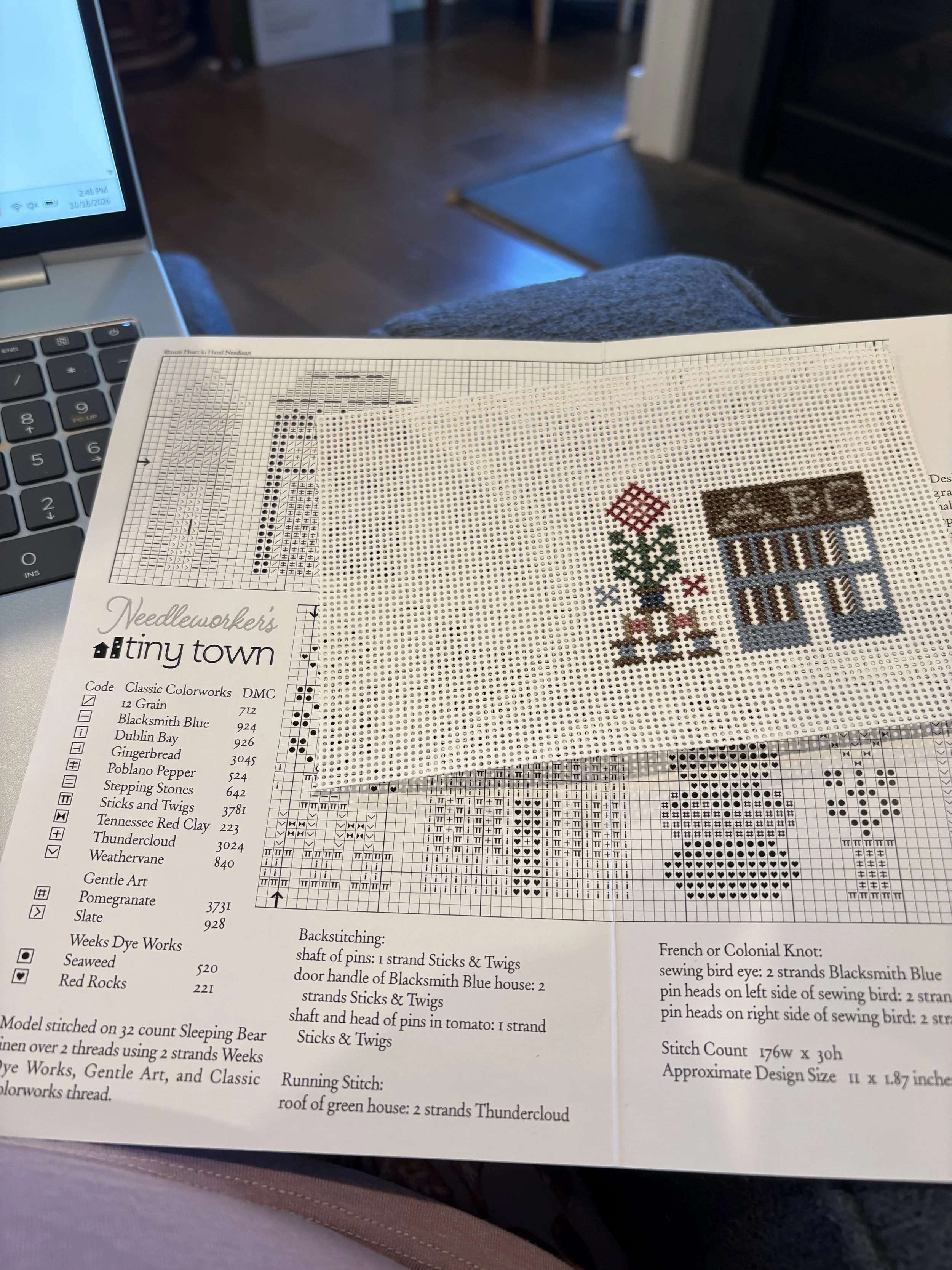 a partially stitched piece on perforated paper shows a little house with part of the alphabet on its roof along with a stylized flower growing from a stack of thread bobbins. beneath the partially stitched item is the pattern from Heart in Hand