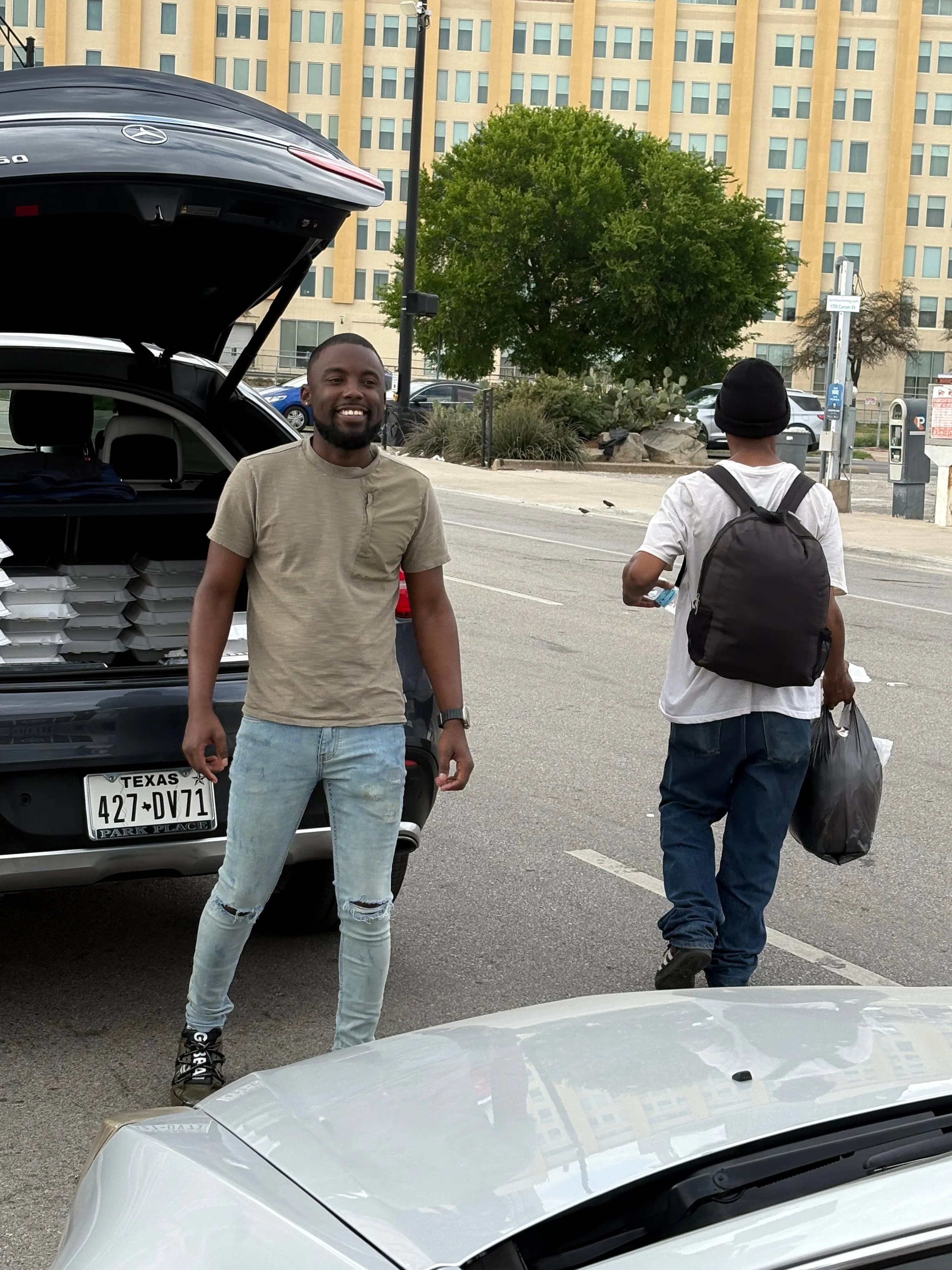 Two men on a city street, one facing the camera smiling, the other walking away with a black backpack and plastic bag, cars parked nearby, a black SUV with open trunk, large building in the background.